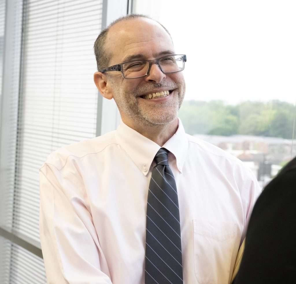 A man with glasses and a beard, wearing a light pink shirt and striped tie, smiling and shaking hands with a woman in a black blazer. They are standing near a window with blinds, and a blurred view of greenery is visible outside.